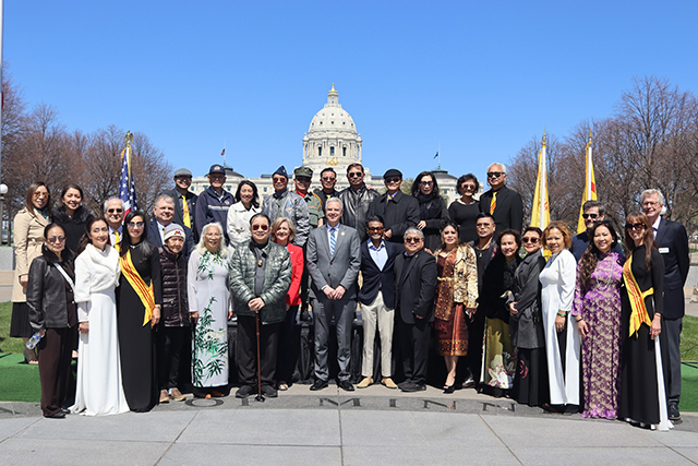 Two rows of people of more than fifteen each stand behind the backdrop of the Minnesota Capitol and a blue sky.